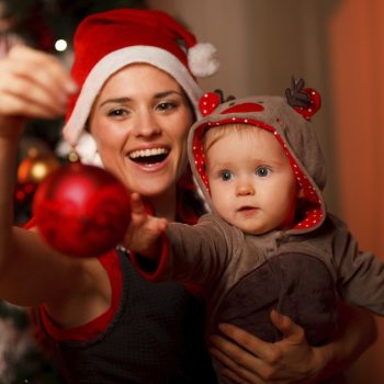 Happy mother showing Christmas ball to baby near Christmas tree Pihenés Szenteste? Eláruljuk, hogy csináld!