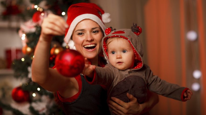 Happy mother showing Christmas ball to baby near Christmas tree Pihenés Szenteste? Eláruljuk, hogy csináld!